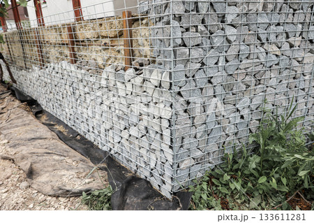 Stone fence in steel mesh and green lawn. Close up of gabion with tightly packed stones of varying sizes and textures. Combination of natural materials and industrial design. Stone fence in steel mesh and green lawn. Close up of gabion with tightly packed stones of varying sizes and textures. Combination of natural materials and industrial design. 133611281
