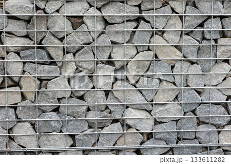 Stone fence in steel mesh and green lawn. Close up of gabion with tightly packed stones of varying sizes and textures. Combination of natural materials and industrial design. Stone fence in steel mesh and green lawn. Close up of gabion with tightly packed stones of varying sizes and textures. Combination of natural materials and industrial design. 133611282