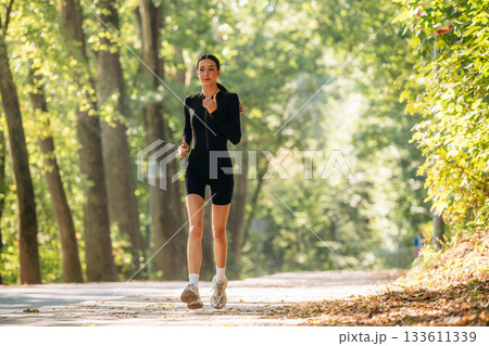 Jogging, front view. Young sportive skinny woman is in black fitness clothes outdoors 133611339