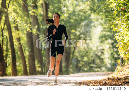 Jogging, front view. Young sportive skinny woman is in black fitness clothes outdoors Jogging, front view. Young sportive skinny woman is in black fitness clothes outdoors 133611340