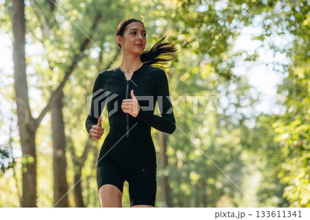 Jogging, front view. Young sportive skinny woman is in black fitness clothes outdoors 133611341
