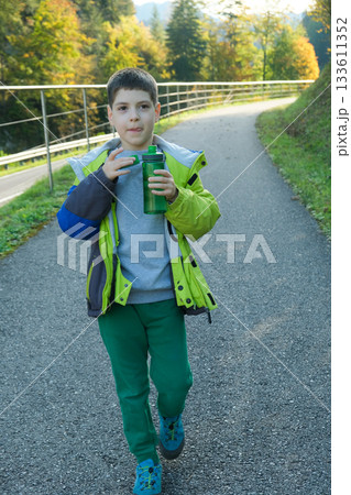A schoolchild walks around the nature and drinks water from a plastic bottle. 133611352