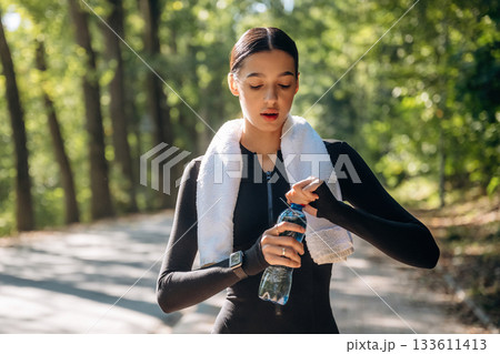 With bottle of water. Young sportive skinny woman is in black fitness clothes outdoors 133611413