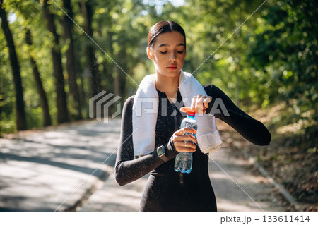 White towel is on the neck. Young sportive skinny woman is in black fitness clothes outdoors 133611414