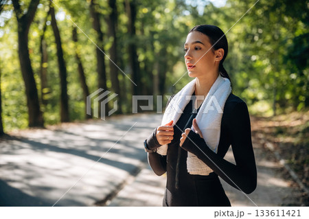 White towel is on the neck. Young sportive skinny woman is in black fitness clothes outdoors 133611421