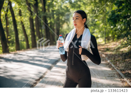 Stay hydrated, with bottle of water, standing. Young sportive skinny woman is in black fitness clothes outdoors Stay hydrated, with bottle of water, standing. Young sportive skinny woman is in black fitness clothes outdoors 133611423