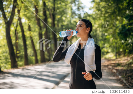 Drinking water. Young sportive skinny woman is in black fitness clothes outdoors Drinking water. Young sportive skinny woman is in black fitness clothes outdoors 133611426
