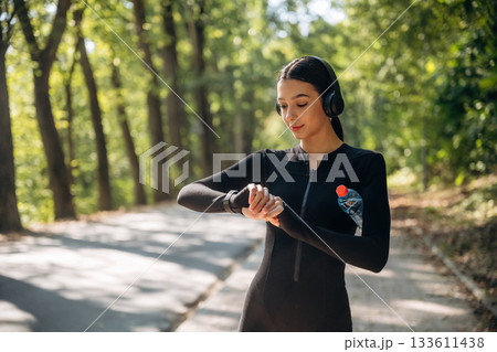 Looking at watch, time, tracker. Young sportive skinny woman is in black fitness clothes outdoors 133611438