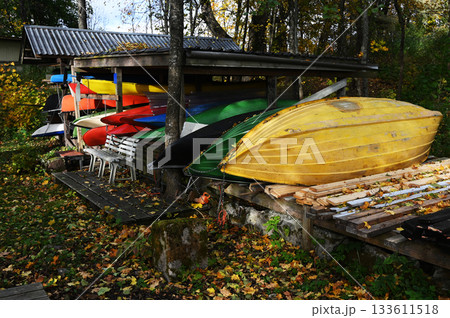 A collection of colorful boats, kayaks, and canoes stored under a wooden shelter in a forested area during autumn 133611518