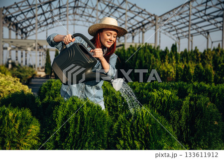 Standing and watering the plants. Young redhead woman is taking care of garden 133611912
