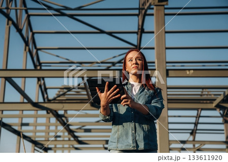 Young redhead woman is standing near unfinished building, construction 133611920