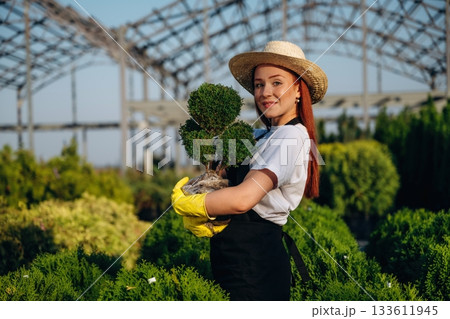 Cheerful facial expression, holding plant. Young redhead woman is taking care of garden 133611945