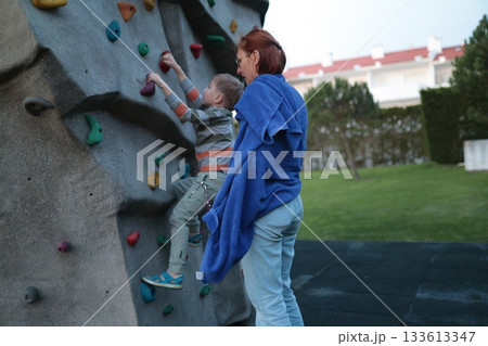 Young boy climbing rock wall with adult support 133613347
