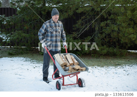 Man with firewood in wheelbarrow and snowy backyard. Winter firewood routine preparing and lighting wood stove. Energy crisis and season concept. 133613506