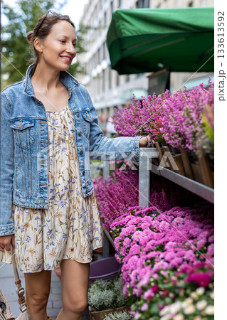 Young adult joyful woman choose to buy colorful potted flowers at outdoor market stall. Female person at sale of blooming plants in pots at botanical street German store on sunny spring autumn day 133613592