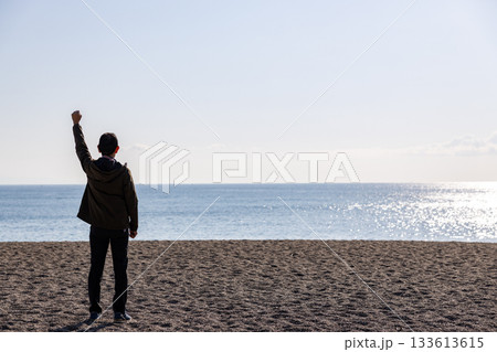 海に向かって片手を上げている一人の男性の後ろ姿 海に向かって片手を上げている一人の男性の後ろ姿 133613615