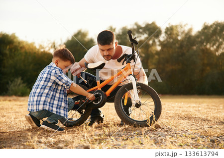 Sitting and repairing the bike. Dad with son are with bicycle on the field outdoors Sitting and repairing the bike. Dad with son are with bicycle on the field outdoors 133613794