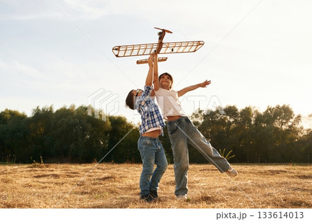 Flying high up. Boy and girl are playing with wooden toy plane on the field Flying high up. Boy and girl are playing with wooden toy plane on the field 133614013