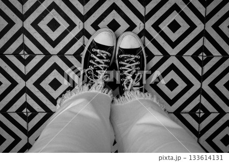 A person's feet and black sneakers are captured from above against a detailed geometric floor. Converse sneakers against a geometric patterned floor. 133614131