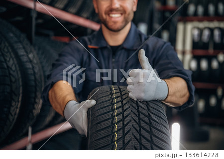 Focused close up view, thumb up, holding a tire. Man worker is maintenance station with wheels 133614228