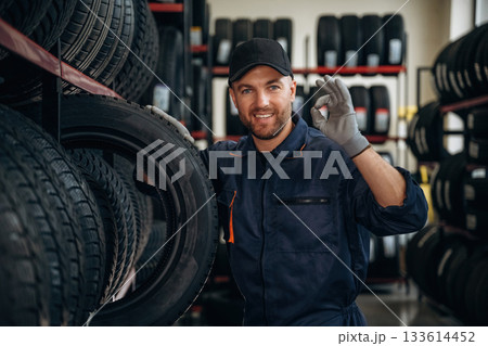 Friendly facial expression, in cap. Man worker is maintenance station with tires, wheels Friendly facial expression, in cap. Man worker is maintenance station with tires, wheels 133614452