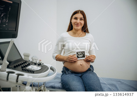 Pregnant woman is sitting in the clinic and holding image of ultrasonic test, conception of anticipation Pregnant woman is sitting in the clinic and holding image of ultrasonic test, conception of anticipation 133614937