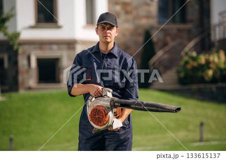 At sunny daytime, using leaf blower. Professional gardener is on the lawn 133615137