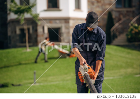 At sunny daytime, using leaf blower. Professional gardener is on the lawn At sunny daytime, using leaf blower. Professional gardener is on the lawn 133615138