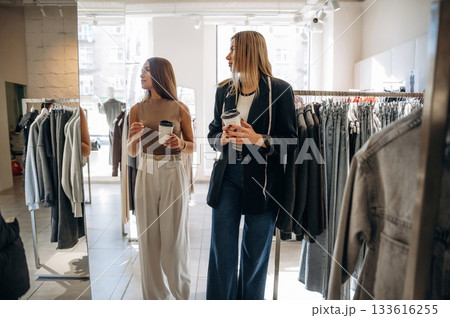 Cups of drinks in hands. Two female friends are in the retail store, choosing clothes, shopping Cups of drinks in hands. Two female friends are in the retail store, choosing clothes, shopping 133616255