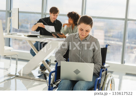 Accessible environment. Woman in chair for people with disability working with laptop in modern office. Diverse multiracial group of people colleagues working together. Young professional team Accessible environment. Woman in chair for people with disability working with laptop in modern office. Diverse multiracial group of people colleagues working together. Young professional team 133617522