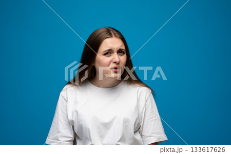 Young woman with confused and skeptical facial expression posing against blue background in studio. 133617626