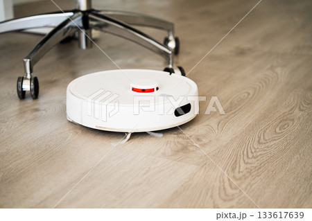 Smart robot vacuum cleaner moving across wooden floor near office chair for efficient automated cleaning 133617639