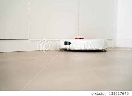 White robotic vacuum cleaning close to wall cabinet, maintaining smooth movement across wooden household flooring White robotic vacuum cleaning close to wall cabinet, maintaining smooth movement across wooden household flooring 133617646