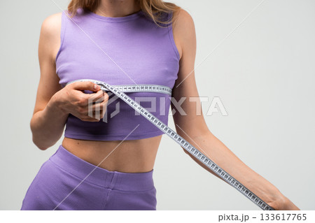Fit woman in purple sports top measuring chest with measuring tape on gray background Fit woman in purple sports top measuring chest with measuring tape on gray background 133617765