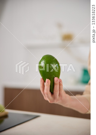 A large avocado in a woman's hand in the interiors of a bright and spacious kitchen A large avocado in a woman's hand in the interiors of a bright and spacious kitchen 133618223