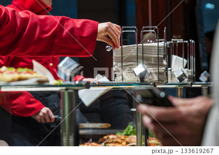 Vendor in a red jacket pulling napkins from a metal dispenser, serving food trays for customers in front. Street food culture and busy market atmosphere. Vendor in a red jacket pulling napkins from a metal dispenser, serving food trays for customers in front. Street food culture and busy market atmosphere. 133619267