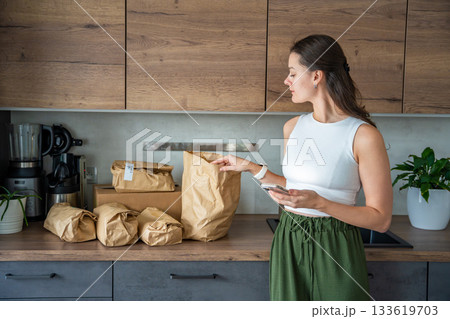 Young woman checks grocery delivery using a mobile app while standing in the kitchen. Concept of app-based meal kit subscription and digital grocery planning for home cooking. Young woman checks grocery delivery using a mobile app while standing in the kitchen. Concept of app-based meal kit subscription and digital grocery planning for home cooking. 133619703