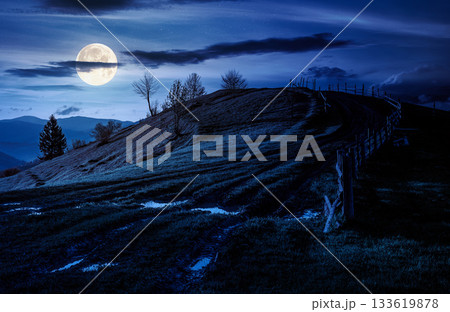wooden fence near the country dirt road through the grassy hill in spring at night. beautiful scenery of mountainous rural area in full moon light. backdrop for fiction art or mystery concepts wooden fence near the country dirt road through the grassy hill in spring at night. beautiful scenery of mountainous rural area in full moon light. backdrop for fiction art or mystery concepts 133619878