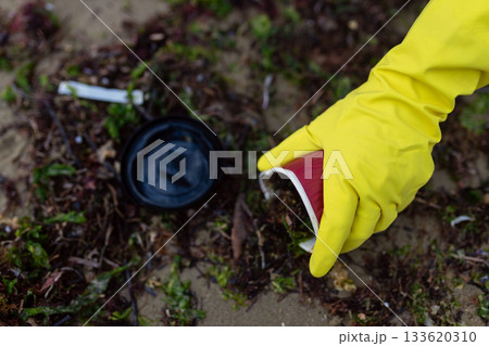 Hands in yellow gloves collecting plastic cup waste during beach cleanup, highlighting pollution, environmental protection and ocean sustainability. Hands in yellow gloves collecting plastic cup waste during beach cleanup, highlighting pollution, environmental protection and ocean sustainability. 133620310