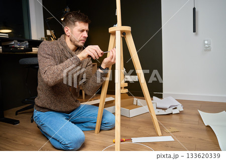 Man kneeling on floor and assembling wooden lamp tripod at home Man kneeling on floor and assembling wooden lamp tripod at home 133620339