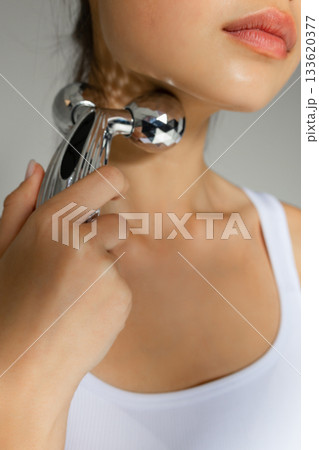 Close-up of a young woman massaging her neck with a metal 3D roller, demonstrating a tightening and relaxing skincare routine at home. 133620377