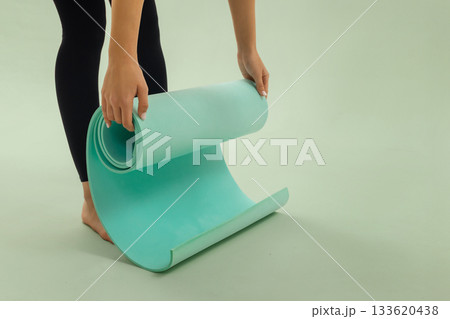 Woman in black leggings unrolls a turquoise yoga mat on a light green background, preparing for workout or stretching session. Woman in black leggings unrolls a turquoise yoga mat on a light green background, preparing for workout or stretching session. 133620438