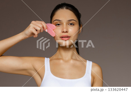 Young woman gently massaging her face with a pink gua sha stone, demonstrating a skincare routine against a neutral background. 133620447