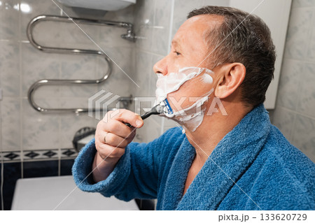 Man in blue robe applies shaving cream and shaves with razor in bathroom. Morning Shaving Routine. Close up. 133620729