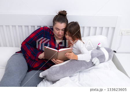 Caucasian female adult reading bedtime story to caucasian female child with stuffed animal on bed 133623605