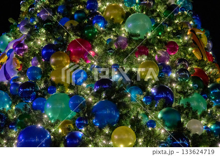 A Christmas tree is packed with glowing lights and glossy ornaments in many colors. The dense branches create a vibrant, festive texture at night. 133624719