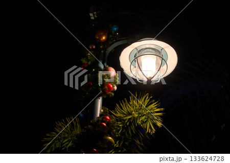 A bright streetlight decorated with Christmas garland and ornaments shines next to palm leaves at night. The roller coaster structure in the background enhances the colorful holiday atmosphere. A bright streetlight decorated with Christmas garland and ornaments shines next to palm leaves at night. The roller coaster structure in the background enhances the colorful holiday atmosphere. 133624728