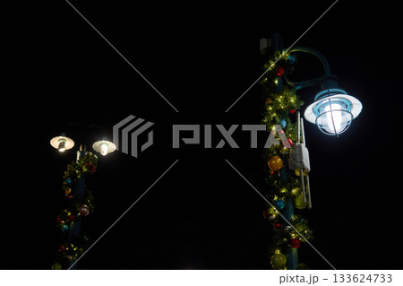 A pair of street lamps decorated with Christmas garland and colorful ornaments glow against a dark sky. The scene shows simple holiday lighting on a quiet walkway in a theme park. 133624733