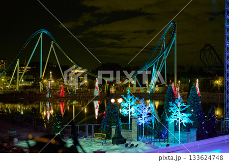 Sweeping roller coaster hills curve above a lagoon lined with glowing holiday trees. Snowy evergreens and show lights add color to the winter night view. Sweeping roller coaster hills curve above a lagoon lined with glowing holiday trees. Snowy evergreens and show lights add color to the winter night view. 133624748
