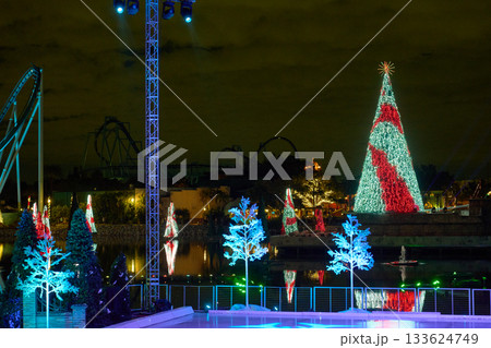 A towering red and white light tree glows across the lagoon next to a bright ice rink. Spotlights, coaster tracks, and floating trees frame the festive scene. A towering red and white light tree glows across the lagoon next to a bright ice rink. Spotlights, coaster tracks, and floating trees frame the festive scene. 133624749